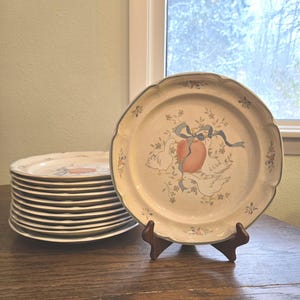 May include: A stack of cream-colored ceramic plates with a scalloped edge and a decorative design featuring doves, a peach, and ribbons. The plates are displayed on a wooden table, with one plate on a wooden stand.