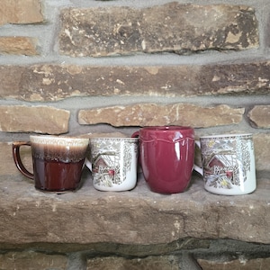 May include: Four mugs and a small pitcher are displayed on a stone shelf. The mugs include a brown and cream-colored mug, two white mugs with a cabin design, and a solid burgundy pitcher. The background is a stone wall.