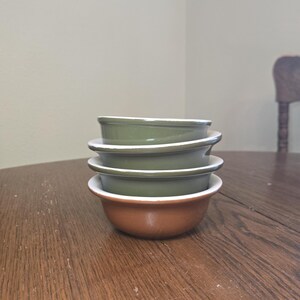 May include: A stack of four ceramic bowls on a wooden table. The bottom bowl is brown, and the three bowls above are olive green with white rims. The bowls are stacked neatly, showcasing their simple, classic design.
