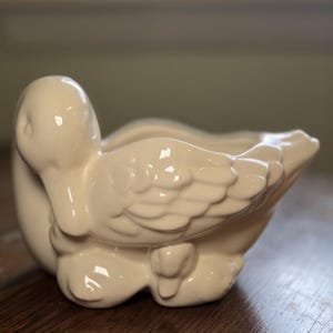May include: Cream-colored ceramic duck planter with a mother duck and two ducklings. The mother duck has detailed wing feathers and a curved neck. The planter is designed to hold small plants or flowers, adding a touch of nature to any space.
