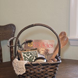 May include: A woven basket filled with baking supplies, including a cookbook, wooden spoons, and decorative tiles. A vintage hand mixer and a lace doily add to the rustic charm. The basket sits on a wooden table.