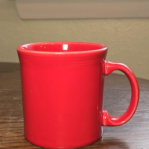 May include: A vibrant red ceramic mug with a rounded body and a curved handle. The mug is glossy and appears to be empty. The mug is sitting on a wooden surface. The mug is a kitchenware item.