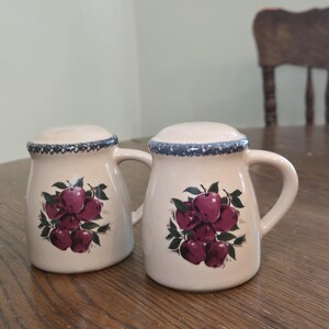 May include: A pair of cream-colored ceramic salt and pepper shakers. Each shaker features a handle and a lid with a blue rim. A cluster of red apples with green leaves decorates the front of each shaker.