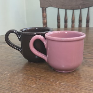 May include: Two ceramic mugs, one dark brown and one dusty rose, sit side-by-side on a wooden surface. Each mug has a rounded body and a curved handle. The background includes a wooden chair.