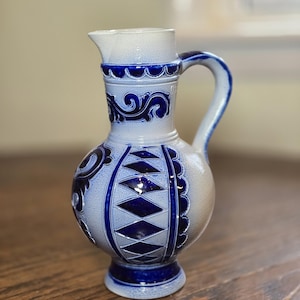 May include: A ceramic pitcher with a bulbous body and a handle. The pitcher is primarily light gray with intricate dark blue patterns, including diamond shapes and scrollwork. It has a spout and sits on a small base.