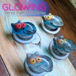 May include: Four "Derpy Tiger" phone grips with a glowing effect. Each grip features a tiger-shaped design with red or yellow eyes and a blue and gray color scheme. The grips are mounted on white and clear bases.