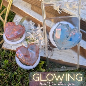 Puede incluir: Tres agarres para teléfono en forma de corazón. Dos son de color rojo-naranja con purpurina, y uno es azul con purpurina. Los agarres están adheridos a bases circulares blancas. El texto "GLOWING Heart Stone Phone Grip" está en la parte inferior.