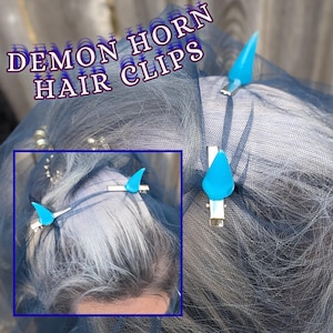 May include: Blue demon horn hair clips. The image shows two blue, horn-shaped hair clips attached to a dark mesh fabric. The clips are silver-toned and the horns are a bright, translucent blue. The text "DEMON HORN HAIR CLIPS" is overlaid on the image.