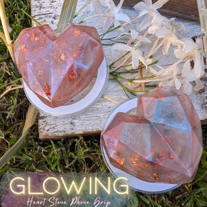 May include: Two heart-shaped phone grips with a faceted design. The grips are a translucent pink with gold flecks and iridescent blue and purple hues. Each grip is mounted on a white circular base. The image includes the text "GLOWING Heart Stone Phone Grip".