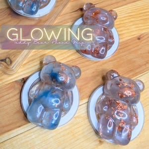 May include: Four glowing teddy bear phone grips. Each grip is a clear resin bear with copper-colored flakes, mounted on a white circular base. The text "GLOWING Teddy Bear Phone Grip" is overlaid on the image.