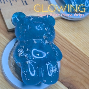 Può includere: Un supporto per telefono a forma di orsetto blu brillante con scaglie d'argento. L'accessorio a forma di orso è realizzato in materiale traslucido e ha una base azzurra. Il testo "GLOWING Teddy Bear Phone Grips" è visibile.