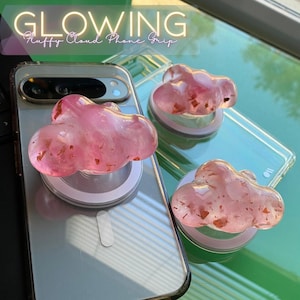 May include: Pink cloud-shaped phone grips with gold flecks are attached to clear phone cases. The phone grips are on a silver phone. The text "GLOWING Fluffy Cloud Phone Grip" is at the top of the image.