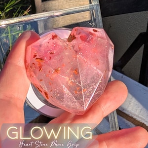 May include: A heart-shaped phone grip with a faceted design. The grip is a translucent pink with gold flecks and the text "GLOWING Heart Stone Phone Grip" at the bottom. The grip is held in a hand.