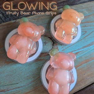 May include: Three glowing, translucent bear-shaped phone grips in shades of peach and pink, each with a green top. The grips are mounted on white and silver circular bases. The text "GLOWING Fruity Bear Phone Grips" is at the top.