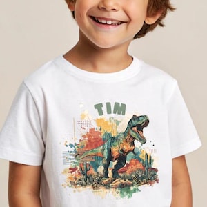 Pode incluir: Camiseta branca com um gráfico colorido de um dinossauro Tyrannosaurus Rex e o nome "TIM". O dinossauro é verde, laranja e azul, com um fundo de paisagem desértica.