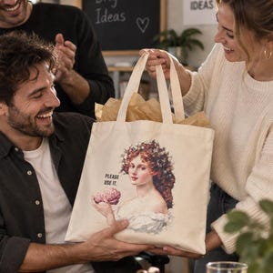 May include: A cream-colored canvas tote bag featuring a printed illustration of a woman with floral hair accessories holding a brain. The text "PLEASE USE IT." is printed in black above the brain. The bag has two sturdy fabric handles.
