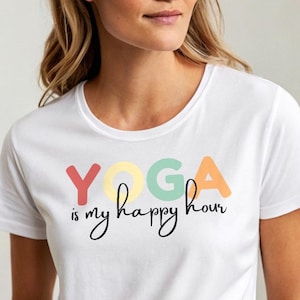 May include: White t-shirt with the text "YOGA is my happy hour" in colorful letters. The word "YOGA" is in red, yellow, green, and orange. The phrase is in a handwritten style font.