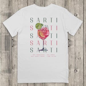 May include: White t-shirt with a cocktail illustration in a stemmed glass. The design includes the word "SARTI" in a vertical pattern, and the ingredients: prosecco, Sarti Rosa, soda, and lime slice.