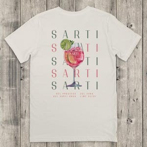 May include: Cream-coloured t-shirt with a cocktail graphic. The design features the word "SARTI" repeated vertically, a cocktail illustration with a lime slice, and the ingredients: Prosecco, Sarti Rosa, Soda, and Lime.