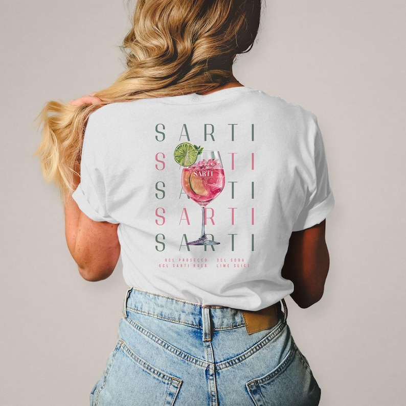 May include: White t-shirt with a cocktail design. The graphic shows the word "SARTI" repeated vertically, with a watercolour illustration of a pink cocktail with a lime wedge. The text "SARTI" is in green and pink.