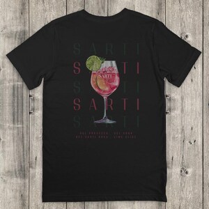 May include: Black t-shirt with a cocktail design. The design includes a stemmed glass with a pink drink, lime garnish, and the word "SARTI" repeated vertically. Ingredients are listed at the bottom.
