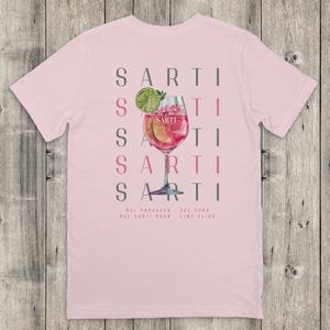 May include: Pale pink t-shirt with a cocktail design. The design features the word "SARTI" repeated vertically, a cocktail glass with a pink drink, and the ingredients: prosecco, Sarti Rosa, soda, and lime slice.