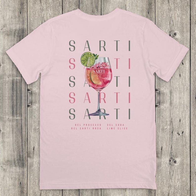 May include: Pale pink t-shirt with a cocktail design. The design features the word "SARTI" repeated vertically, a cocktail glass with a pink drink, and the ingredients: prosecco, Sarti Rosa, soda, and lime slice.