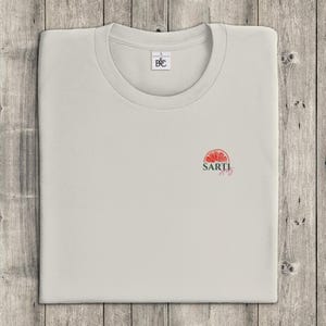 May include: A pale grey t-shirt with a crew neck, neatly folded on a wooden surface. The shirt displays a small, red and green graphic of a citrus fruit with the word "SARTI" beneath it. A small tag is near the collar.