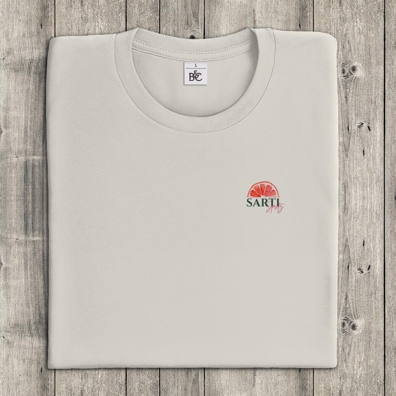 May include: A pale grey t-shirt with a crew neck, neatly folded on a wooden surface. The shirt displays a small, red and green graphic of a citrus fruit with the word "SARTI" beneath it. A small tag is near the collar.