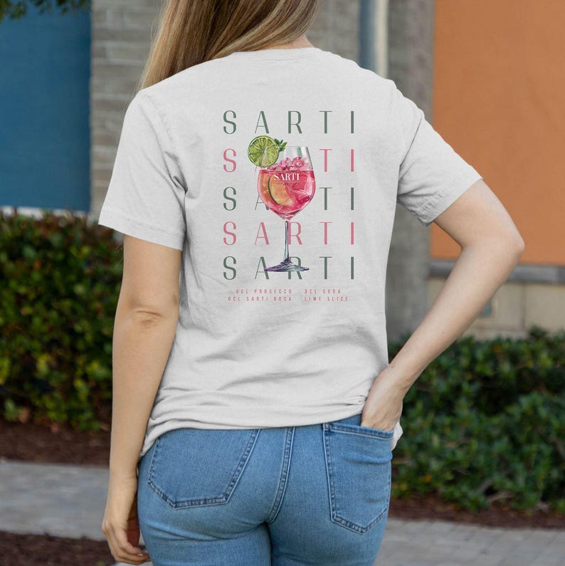 May include: A pale grey t-shirt with a cocktail graphic in a wine glass, the word "SARTI" repeated vertically. The cocktail is pink with a lime garnish. Below the graphic are the words "Sui Prosecco, Del 1984, Sui Sarti Rosa, Lime Slice."
