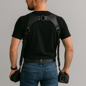 Puede incluir: Arnés de cámara de cuero negro con correas ajustables y herrajes metálicos. El arnés está diseñado para sujetar dos cámaras, con puntos de fijación en el pecho y los lados. Se lleva sobre una camiseta negra y vaqueros azules.