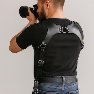 Puede incluir: Arnés de cámara de cuero negro con herrajes metálicos plateados. El arnés está diseñado para llevarse sobre los hombros y la espalda, con correas y hebillas ajustables. Está diseñado para sujetar una cámara.