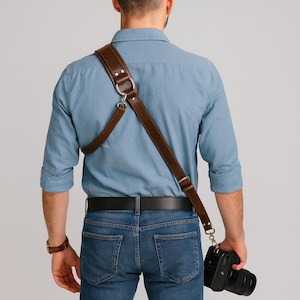 May include: A brown leather camera strap with silver metal hardware. The strap is worn over the shoulder and connects to a black camera. The person is wearing a blue shirt and jeans.