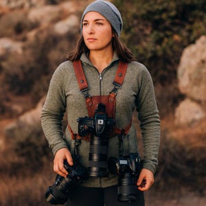 May include: A brown leather camera harness with two cameras attached. The harness has adjustable straps and a chest plate. The person in the image is wearing a grey beanie and a green zip-up jacket.