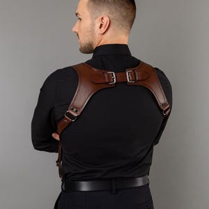 May include: A brown leather harness with adjustable buckles and straps. The harness is worn over a black shirt. The leather is a rich, dark brown colour. The straps cross the shoulders and chest, with metal hardware.