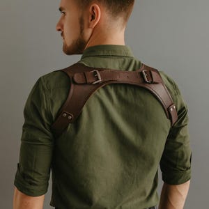 May include: A brown leather shoulder harness with metal buckles and rivets. The harness is worn over a green button-down shirt. The leather appears to be distressed, and the buckles are silver-coloured.