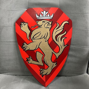Puede incluir: Un escudo de madera en forma de escudo con un diseño rojo y dorado. El escudo presenta un león dorado rampante con una corona, sobre un fondo rojo con rayas diagonales. El emblema se muestra sobre una superficie gris.
