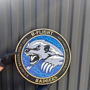 May include: A circular wall plaque featuring a snarling grey badger graphic on a blue background with lightning bolt accents. The black border contains the gold text B-FLIGHT BADGERS DON'T GIVE.....ANYTHING BUT EXCELLENCE!