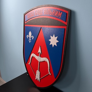 Puede incluir: Un escudo de madera con un centro rojo, detalles azules y borde negro. El escudo presenta un arco y una flecha, una flor de lis y una estrella. El texto "НЕБО БУДЕ ВІЛЬНИМ" se muestra en la parte superior en rojo.
