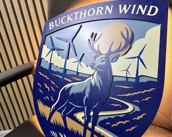 Könnte beinhalten: Ein mehrschichtiges Holzschild mit dem Text "BUCKTHORN WIND" oben. Das Schild zeigt einen blauen Hirsch, Windräder und ein Landschaftsdesign in Blau-, Beige- und Gelbtönen. Das Schild hat die Form eines Schildes.