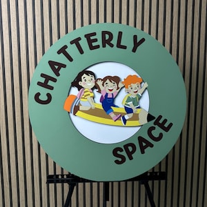 Puede incluir: Un letrero redondo verde con las palabras "CHATTERLY SPACE" en letras marrones. El letrero presenta un interior blanco con tres niños de dibujos animados montados en un lápiz. El letrero está montado sobre un soporte negro.