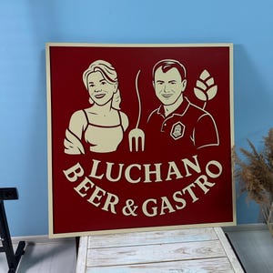 Puede incluir: Un letrero cuadrado de madera con fondo burdeos y borde color crema. El letrero presenta ilustraciones de un hombre y una mujer, un tenedor y las palabras "LUCHAN BEER & GASTRO" en letras color crema. El letrero es probablemente para un restaurante o bar.