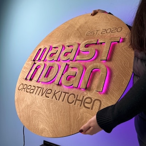 Puede incluir: Un letrero de madera con las palabras "Maast Indian Creative Kitchen" y "Est. 2020" grabadas. Las letras están iluminadas con un brillo rosa, creando un contraste llamativo con la madera marrón claro.