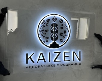 Könnte beinhalten: Rechteckiges, beleuchtetes Schild mit dem Wort "KAIZEN" in fetter, serifenloser Schrift. Das Schild zeigt ein kreisförmiges Logo mit Federdesign und botanischen Elementen. Der Text "АДВОКАТСЬКЕ ОБ'ЄДНАННЯ" steht darunter.