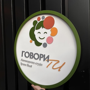 Może przedstawiać: Okrągły znak z białym tłem i zieloną obwódką. Znak przedstawia kreskówkową ilustrację uśmiechniętej twarzy z kolorowymi kształtami i tekstem w obcym języku. Tekst zawiera słowa "ГОВОРИ" i "ти".
