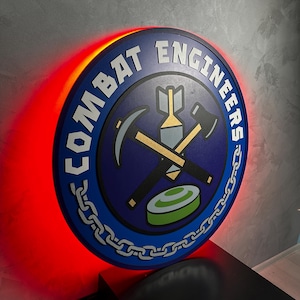 May include: A round sign with the words "COMBAT ENGINEERS" in white letters. The sign has a blue background with a central emblem featuring crossed tools and a bomb. A red light illuminates the back of the sign.