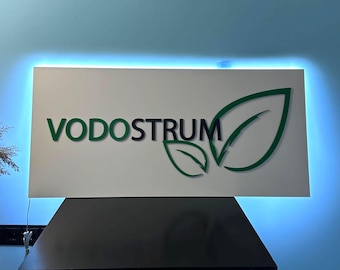 Könnte beinhalten: Ein rechteckiges weißes Schild mit dem grünen Wort "VODOSTRUM" und einem grünen Blattlogo. Das Schild hat einen blau beleuchteten Rand und ist auf einer schwarzen Oberfläche montiert.