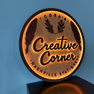 以下が含まれることがあります： 温かい光で照らされた、筆記体フォントで「Creative Corner」の文字が入った丸い木製の看板。看板には「Nashville Stationery」の文字と装飾用の羽と葉も付いています。看板は茶色で、中央は明るい木材です。