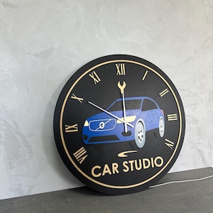 Peut inclure: Une horloge murale ronde noire avec une bordure dorée et des chiffres romains. Le cadran de l'horloge présente un dessin de voiture bleue et les mots "CAR STUDIO". Les aiguilles sont dorées.