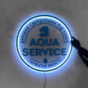 Puede incluir: Un letrero redondo e iluminado con el texto "AQUA SERVICE" en azul. El letrero tiene un borde azul brillante y texto en ucraniano. Una mano con un guante negro está tocando el letrero.